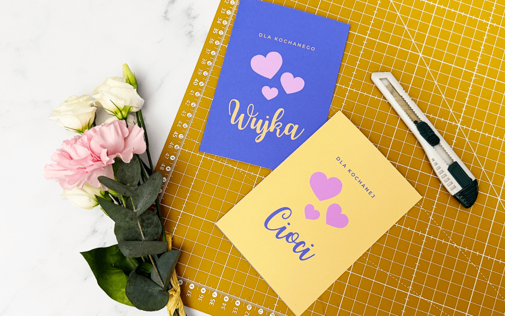 Dwie kartki - jedna żółta dla kochanej Cioci, druga fioletowa dla kochanego Wujka, leżą na żółtej macie do wycinania. obok nożyk do papieru i bukiet kwiatów.