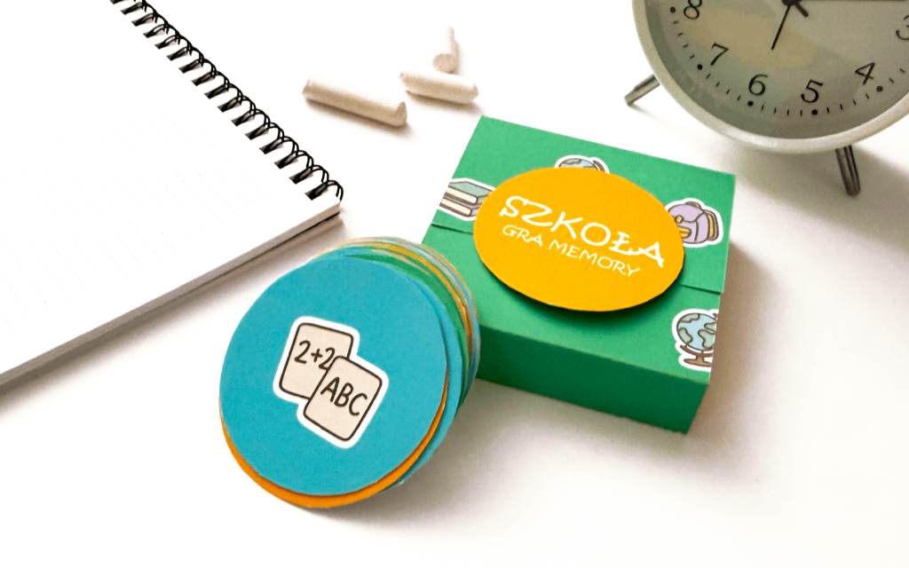 Pudełko z napisem SZKOŁA gra memory - widok z góry na tle jasnego biurka z kredą i zegarem.