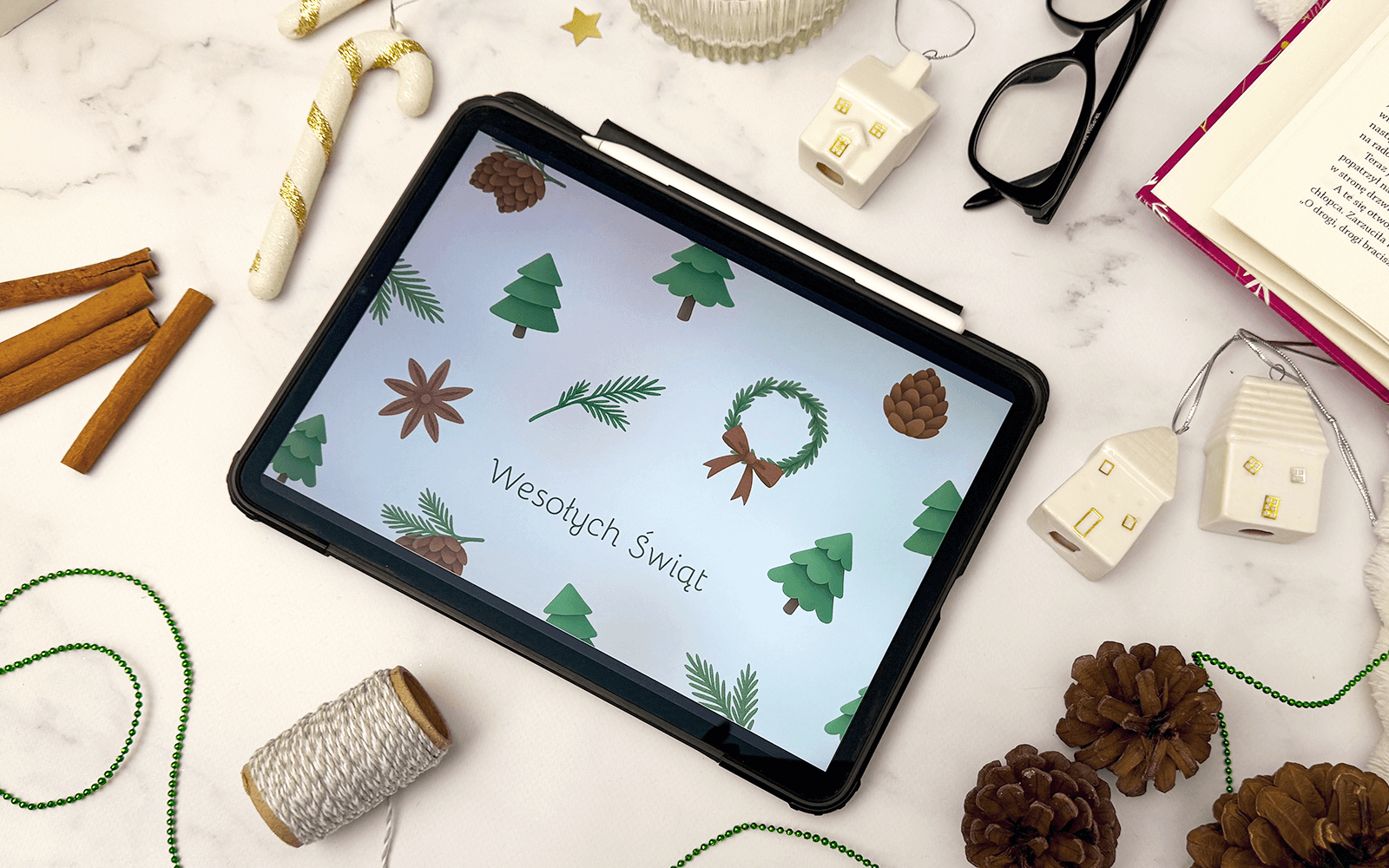 Kartka świąteczna w wersji cyfrowej wyświetlona na tablecie. Wokół akcenty świąteczne, takie jak dekoracje choinkowe, cynamon i szyszki.