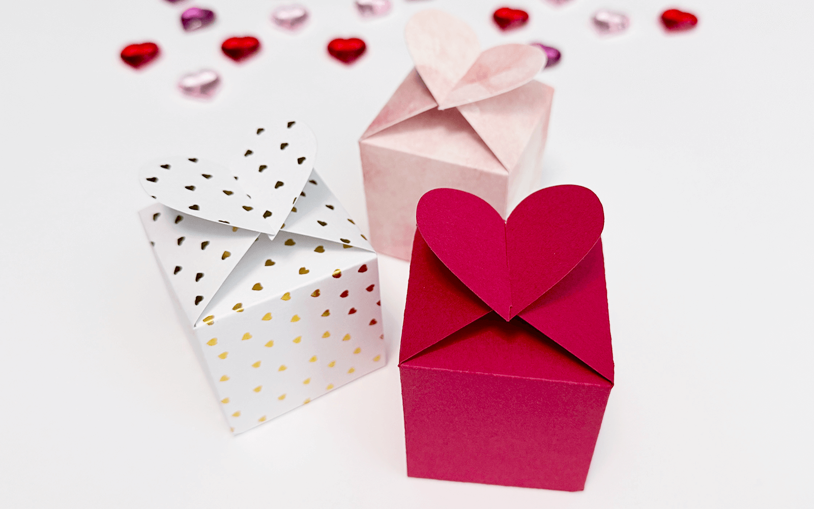 Trzy papierowe pudełeczka DIY z wieczkiem w kształcie serduszka. Wszystkie trzy zostały złożone z odmiennych papierów ozdobnych - białego w złote serduszka, różowego z akwarelowym motywem i czerwonego.